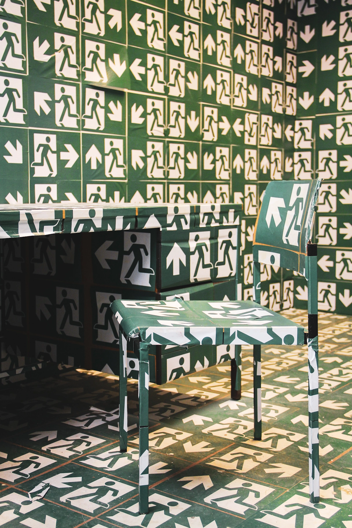 Ein Raum im Kunstlabor mit einem Tisch und einem Stuhl. Beides ist inklusive Wand und Boden mit hunderten Notausgangschildern beklebt