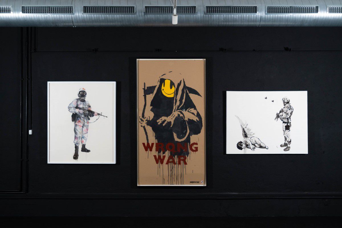3 painted pictures in the MUCA museum, on the left a man with gas mask and rifle, in the middle a grim reaper with smiley as face and the inscription 
