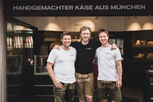 Die drei Besitzer der Münchner Käse Manufaktur stehen vor ihrem Laden. Im Hintergrund sind Regale mit Käselaibe zu entdecken