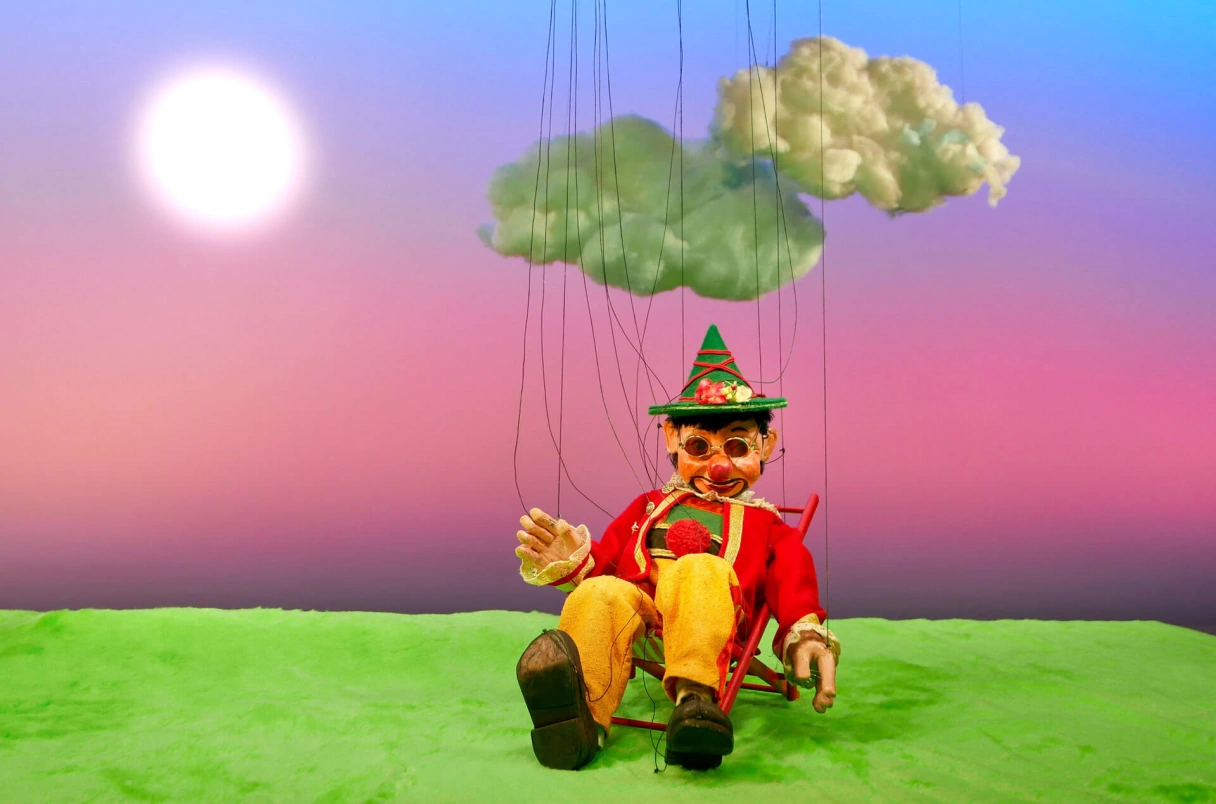 Marionette bunt gekleidet mit Hut unter zwei Wolken sitzend, auf einer Wiese mit lila-blauem Hintergrund