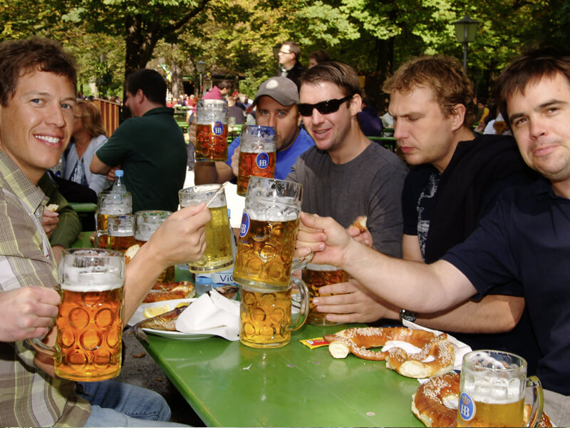 Eine Gruppe im Biergarten am Tisch mit jeweils einer Maß in der Hand und Brezeln auf dem Tisch