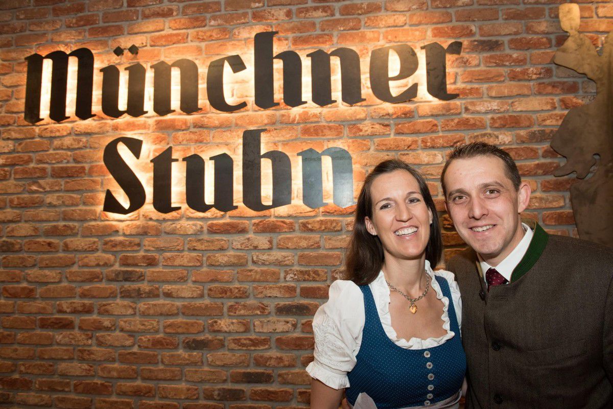 Mrs. and Mr. Wickenhäuser, the owners of the Münchner Stubn, in front of a stone wall with a glowing 