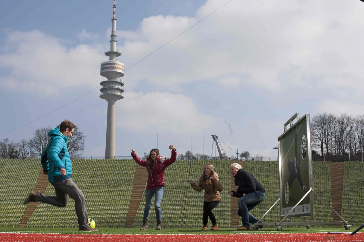 Aufgebaute Torwand im Olympiastadion München mit Olympiaturm im Hintergrund. Ein Mann schießt den Ball zur Torwand, 3 Zuschauer jubeln