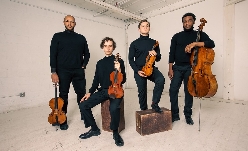 4 young men pose with their string instruments. They are all dressed in black