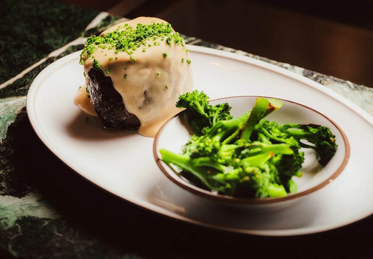 An elegantly plated dish featuring a piece of meat smothered in a creamy sauce and sprinkled with fresh herbs, served on a white plate alongside green broccoli, in the warm glow of a kitchen.