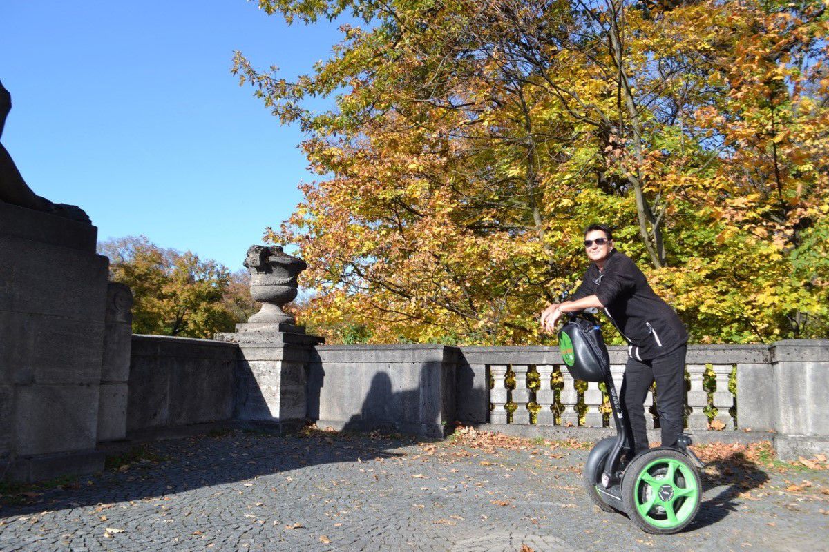 Seg to rent München, Tourist in schwarzer Kleidung steht auf einem Segway mit grünen Rädern, Herbstliche Stimmung mit einem bunten Laubbaum im Hintergrund