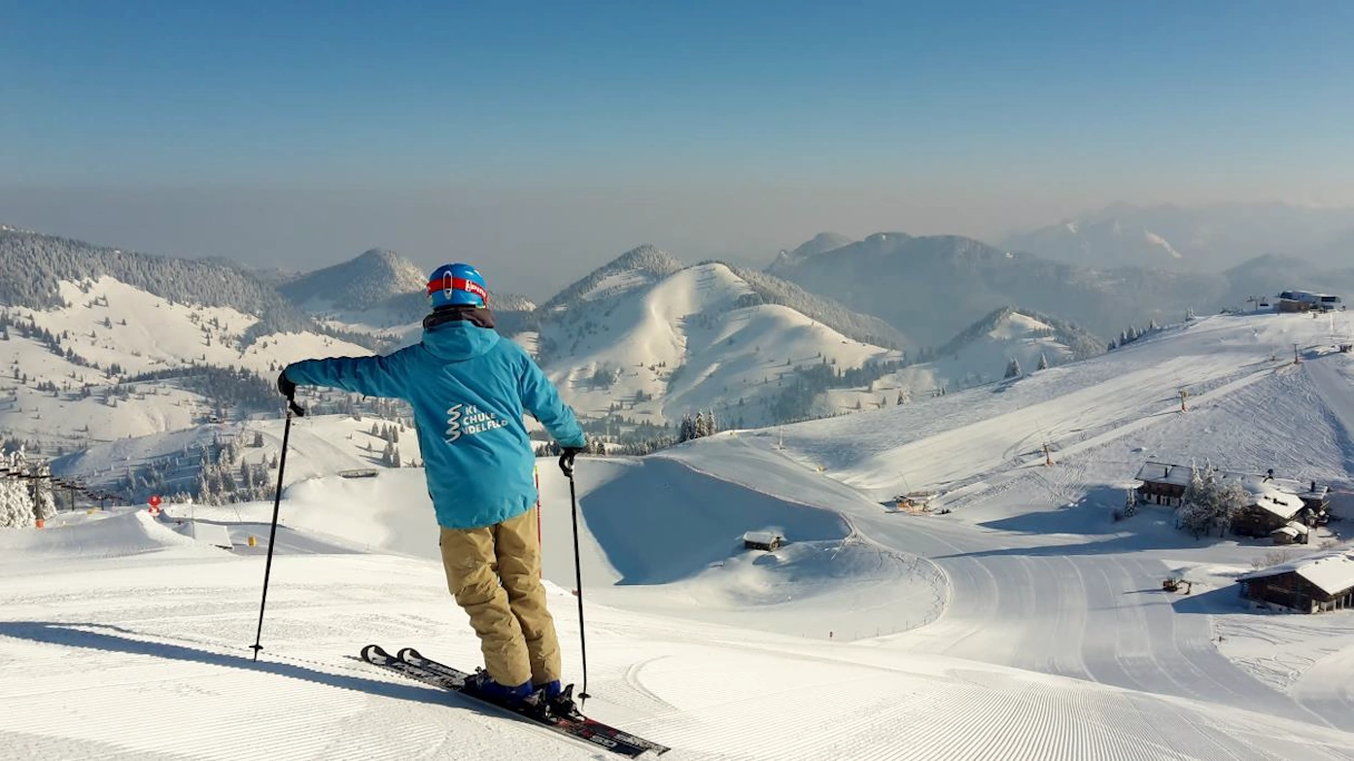 Junger Mann auf Skiern steht auf einem Berg in Sudelfeld. Im Hintergund sind schneebedeckte Berge zu sehen