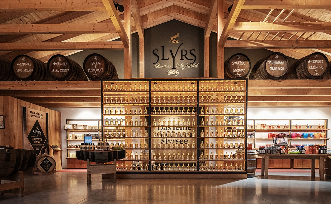 Slyrs Whisky Shop mit vielen edlen Flaschen und Produkten zum Verkaufen aufgereiht. Holzfässer und SLYRS Logo fallen als Deko auf