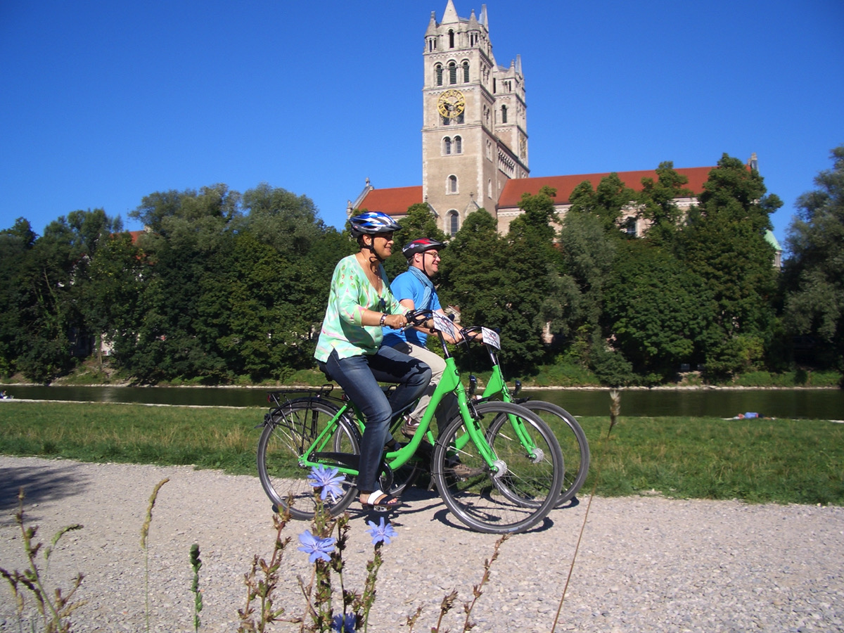 Spurwechsel Fahrradverleih, Fahrradfahrer fahren an der Isar entlang, im Hintergrund ist eine große Kirche zu sehen