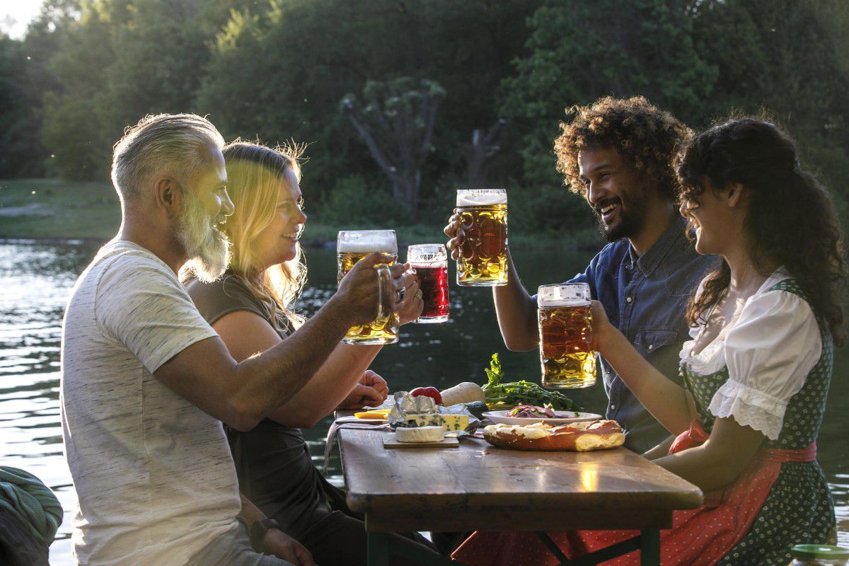 Biergarten am Wasser, mit 4 Gästen, die mit einer Maß Bier anstoßen und bayerischem Essen auf dem Tisch