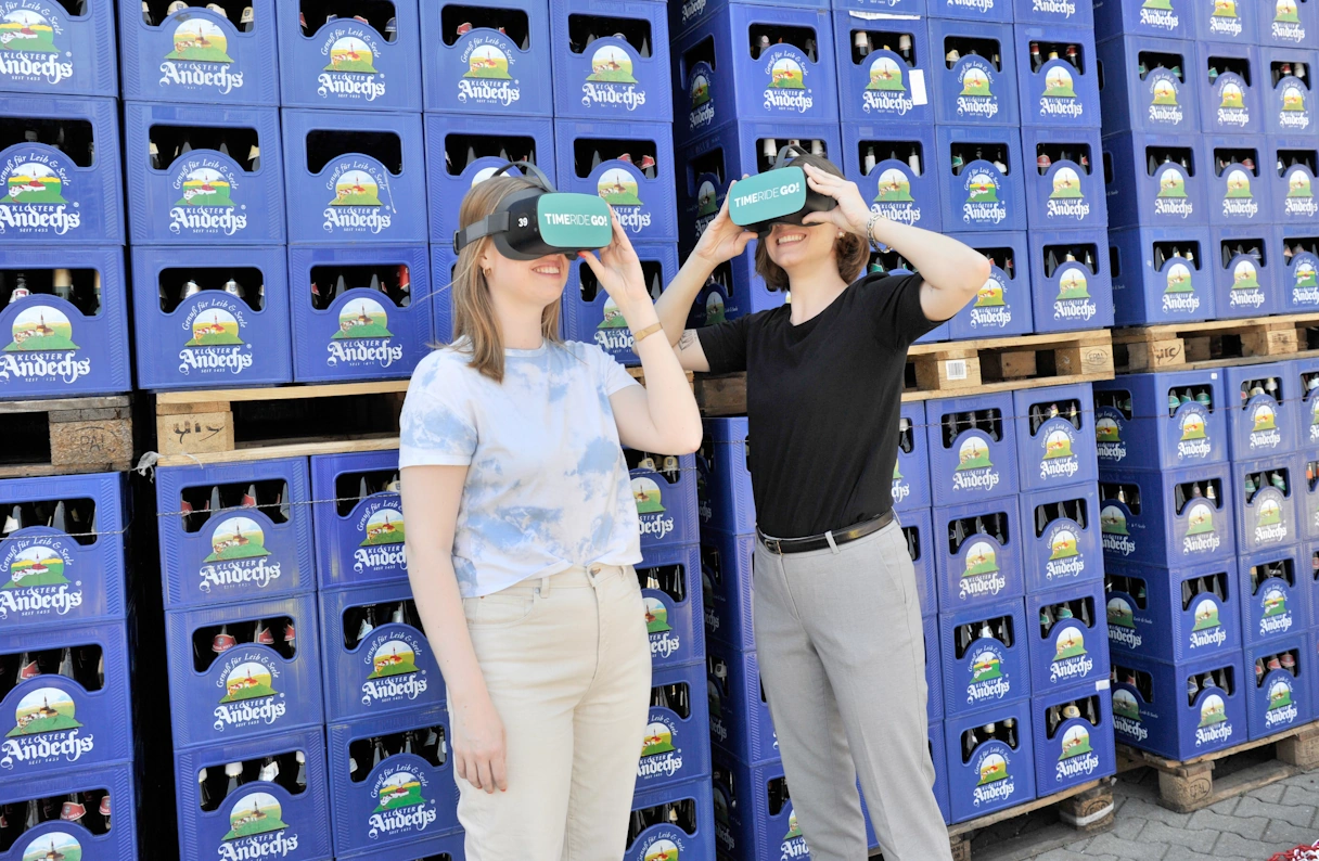 Zwei junge Frauen mit der TimeRide GO! VR Brille vor gestapelten Bierkästen