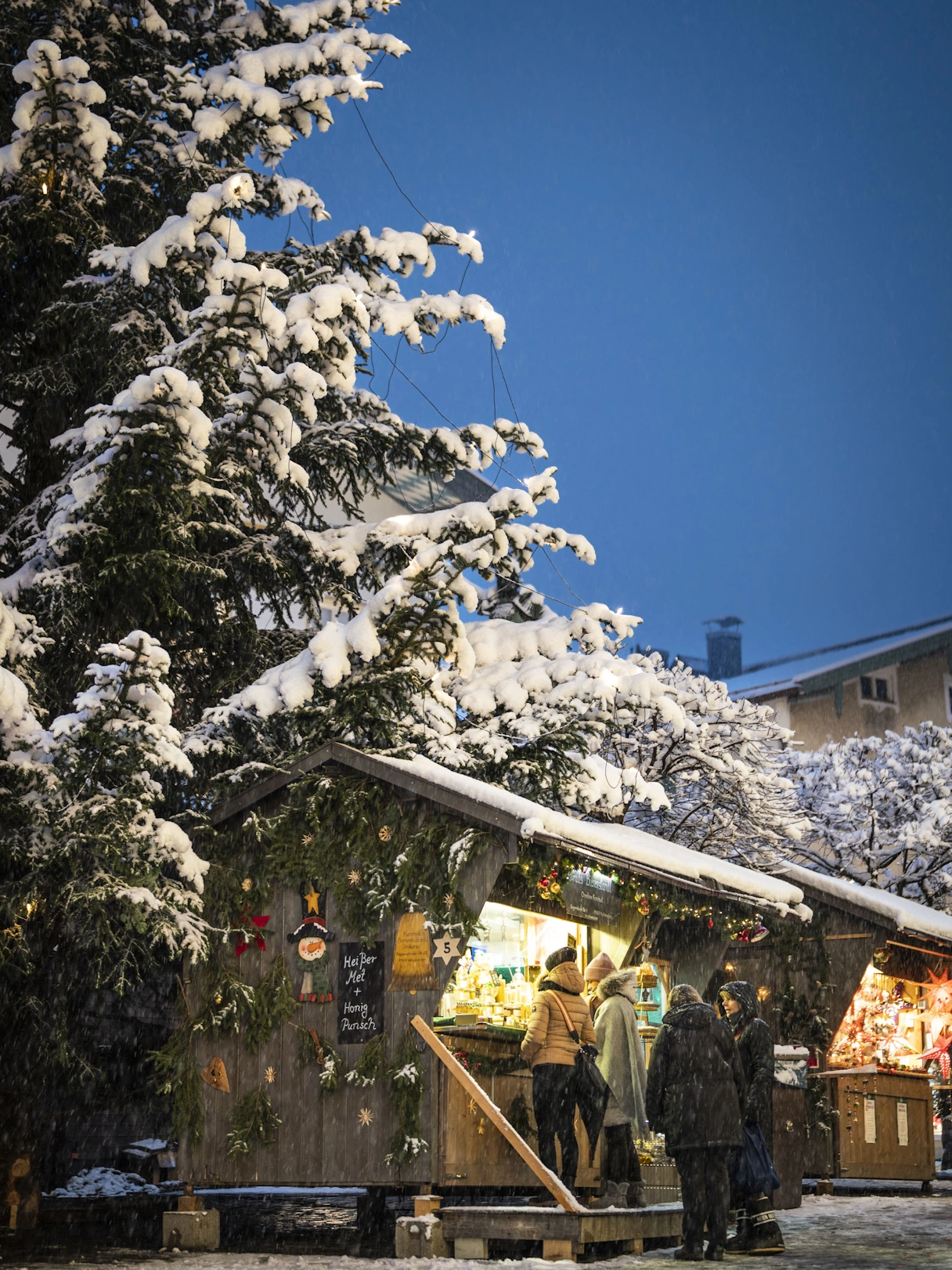Große Tanne mit Schnee bedeckt, darunter ein Christkindlmarkt Stand