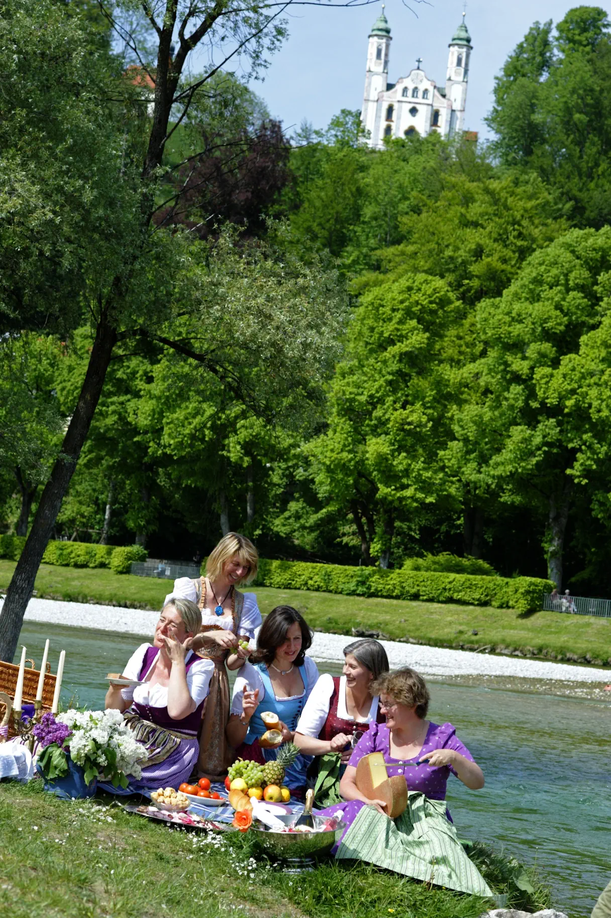 Die 5 Tölzer Stadtversucherinnen im Dirndl gekleidet sitzen auf der Wiese vor der Isar und picknicken.