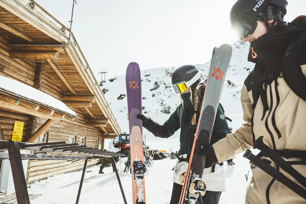 Two people in winter sports equipment stand in front of a hut with skis and are surrounded by snow