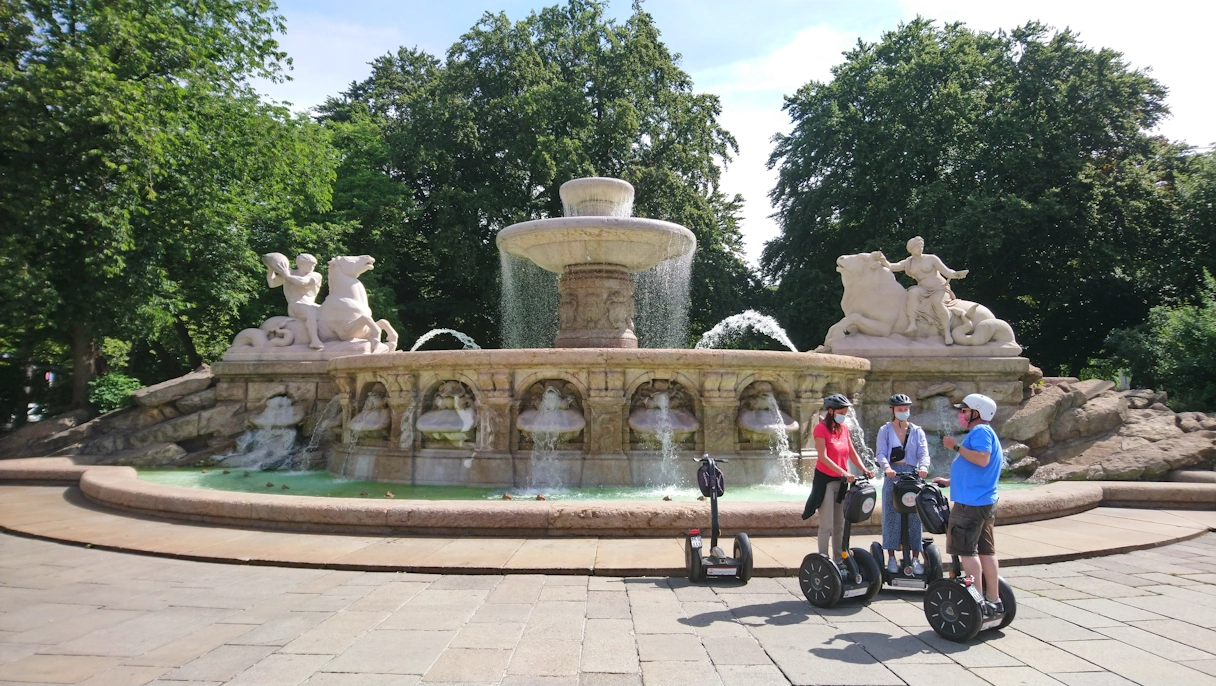 Segway Tour zu dritt in München im Sommer. Die drei Personen stehen vor einem großen Brunnen