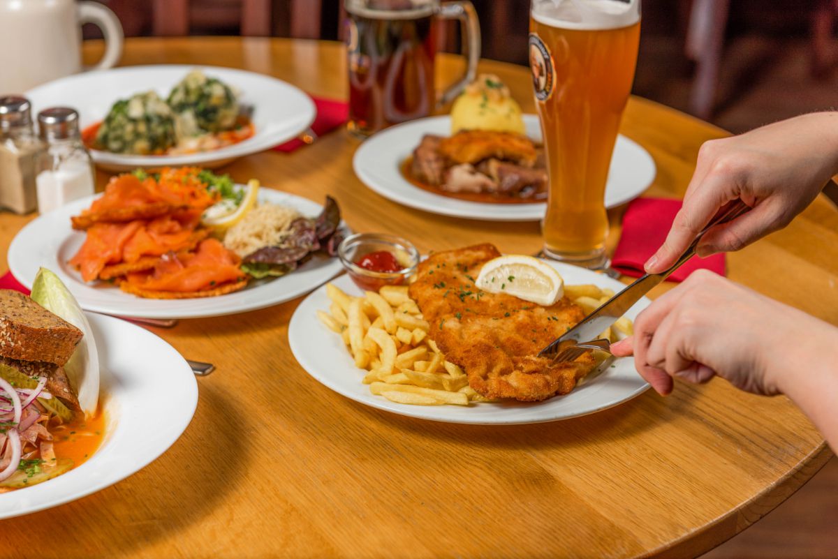 Wirtshaus im Schlachthof, München, Tisch mit fünf vollen Tellern und verschiedenen Getränken stehen auf dem Tisch, Schnitzel mit Pommes wird gerade angeschnitten
