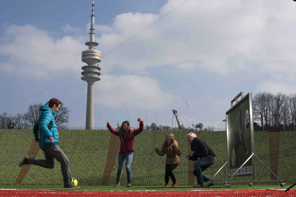 vier Personen spielen Fußball vor einer Fußballwand, im Hintergrund steht der Olympiaturm