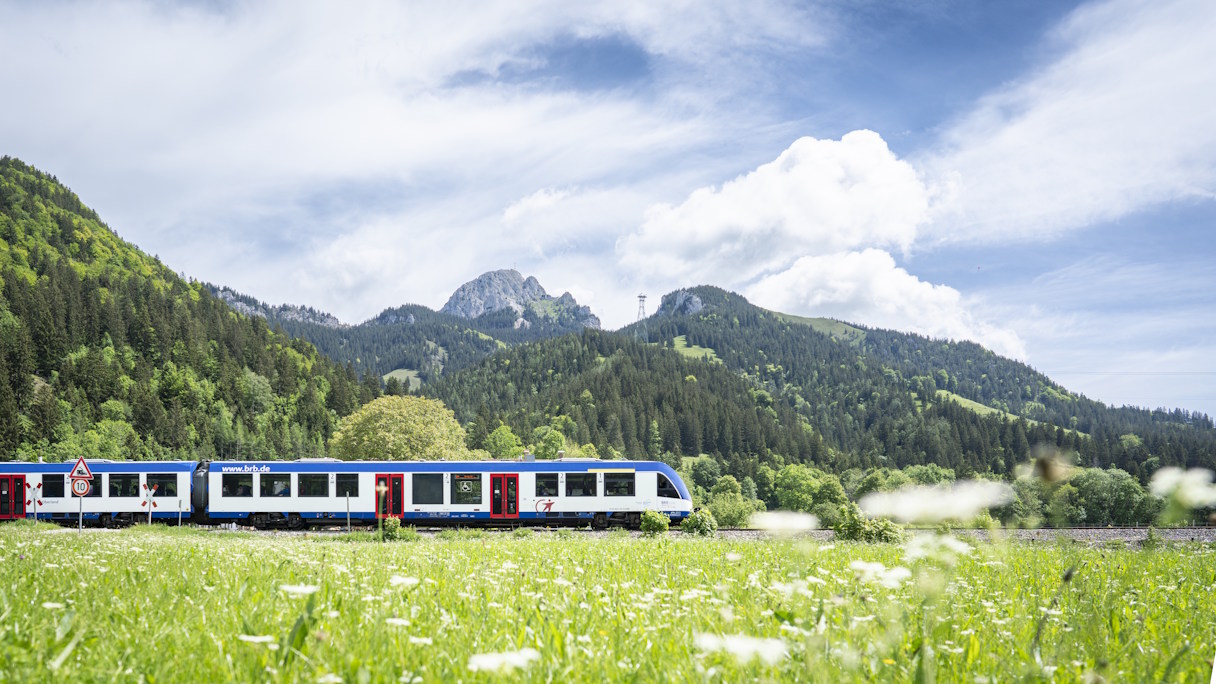 Zug fährt durch Landschaft bei Geitau, umgeben von Bäumen und Bergen.